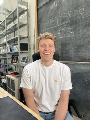Smiling man in a white shirt sits in front of a chalkboard with shelves of books nearby.