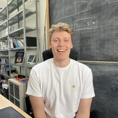 Smiling man in a white shirt sits in front of a chalkboard with shelves of books nearby.