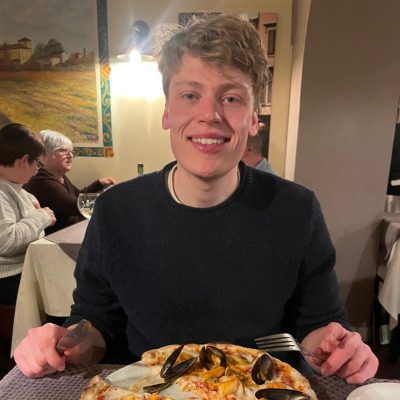 A person smiling at a restaurant table, holding cutlery, with a plate of seafood pizza in front. Other diners are in the background.