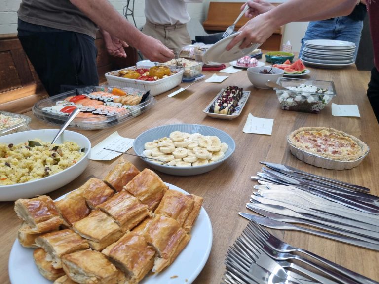 A buffet table with various dishes, including pastries, sushi, fruit, quiche, and desserts. People are serving themselves.