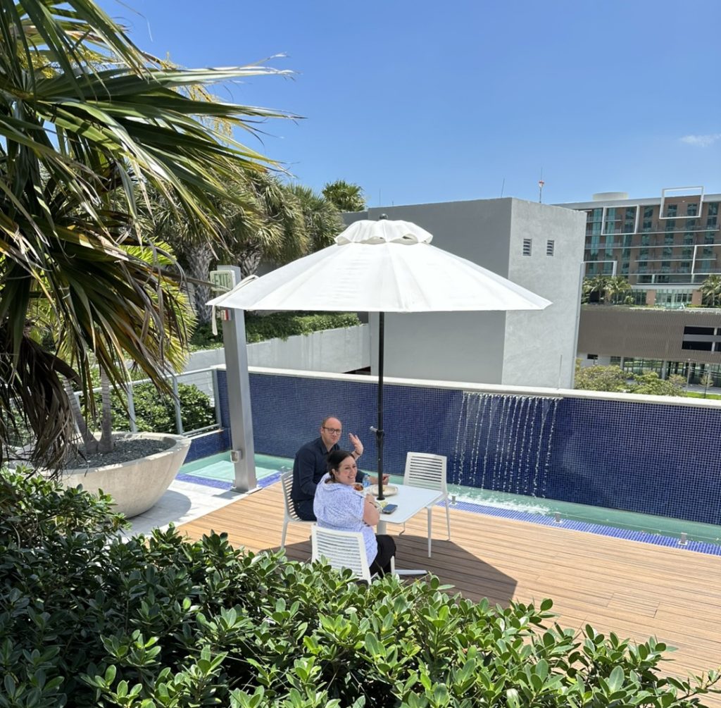 Two people sit under a white umbrella by a pool with a cascading waterfall, surrounded by greenery, on a sunny day.