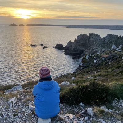 Person in a blue jacket and red hat sits on a rocky cliff, overlooking a coastal landscape at sunset, with calm water and cliffs.