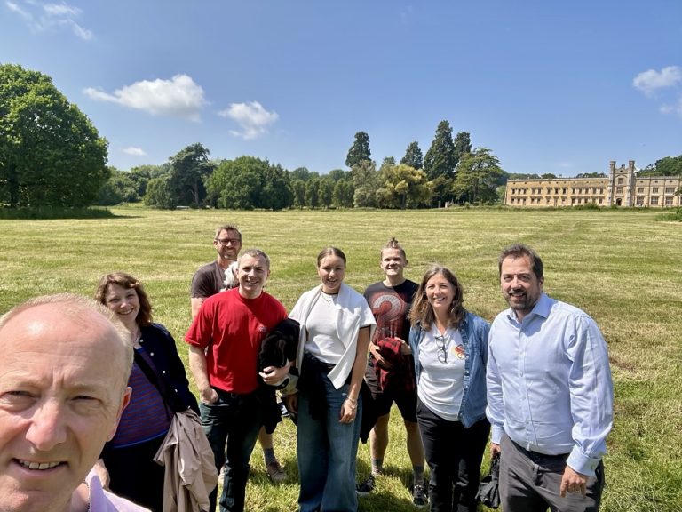 A group of people standing on a sunny, grassy field with trees and a large building in the background.