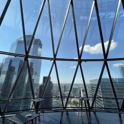 View of a city skyline through the geometric, glass-paneled windows of a modern skyscraper, with tables and chairs inside.