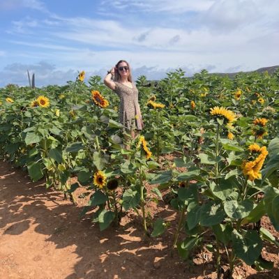 A person stands in a sunflower field on a sunny day, wearing sunglasses and a patterned dress, with a clear blue sky overhead.