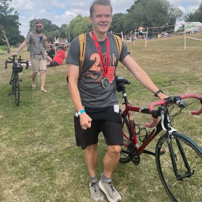 A person stands smiling next to a red bicycle, wearing medals and a race t-shirt in a grassy park with people and trees around.