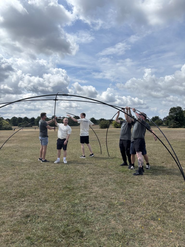 Five people assembling a large black metal framework on a grassy field under a partly cloudy sky.
