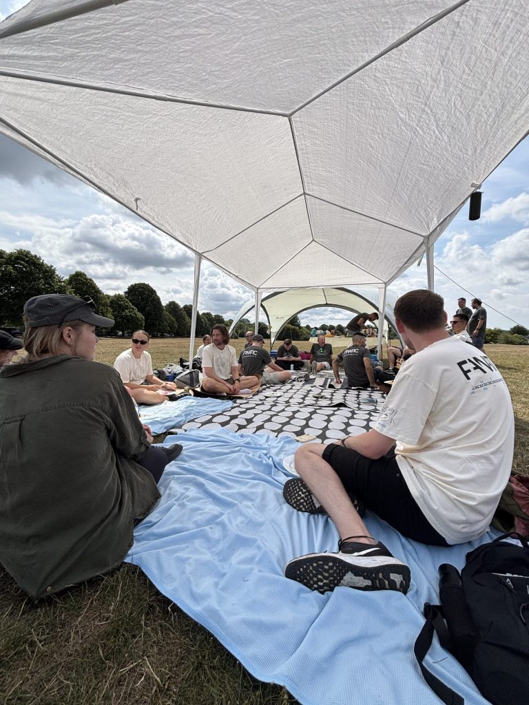 A group of people sitting under a white canopy on patterned and blue blankets in an open grassy area, enjoying a casual outdoor gathering.