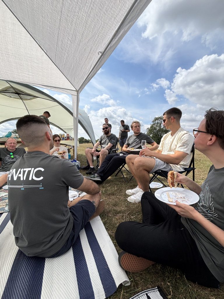 A group of people sitting under a tent at a picnic, eating and talking. The sky is partly cloudy.