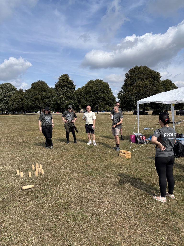 People standing in a park playing a lawn game with wooden pins and batons, under a partly cloudy sky near trees and a tent.