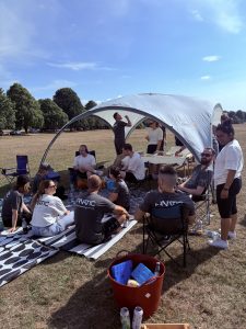 A group of people relax under a canopy in a park on a sunny day. Some are sitting on rugs or chairs, and a table has refreshments.