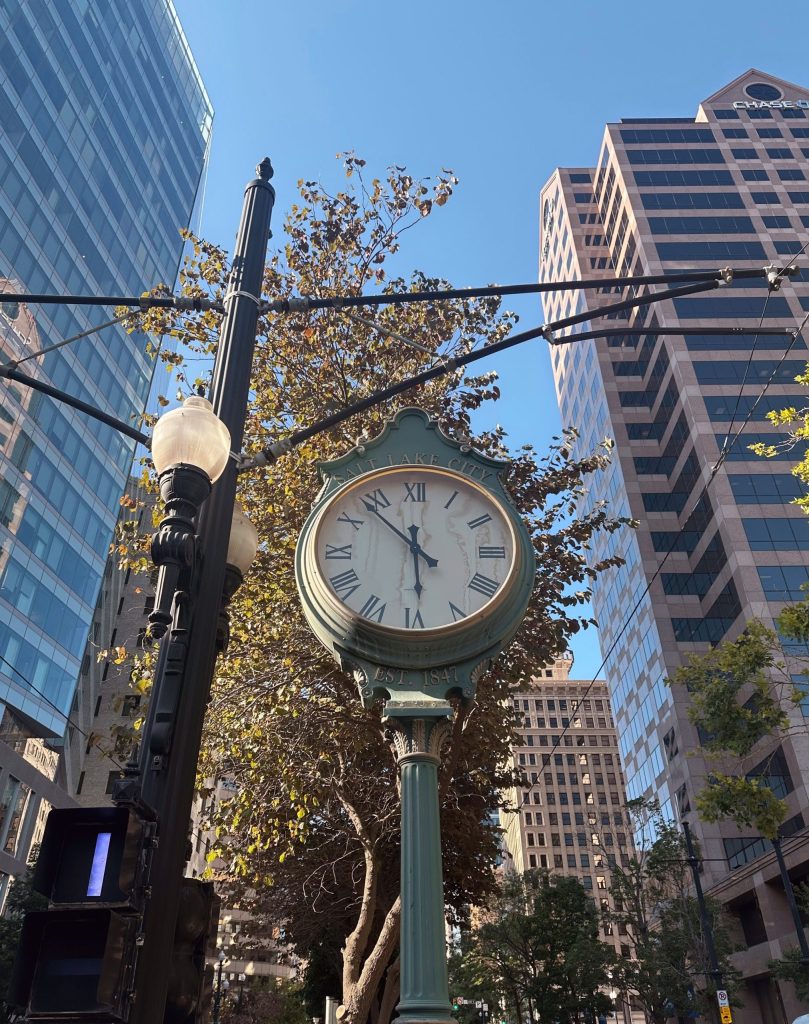 A vintage street clock with Roman numerals stands in front of tall modern buildings and trees, under a clear blue sky.