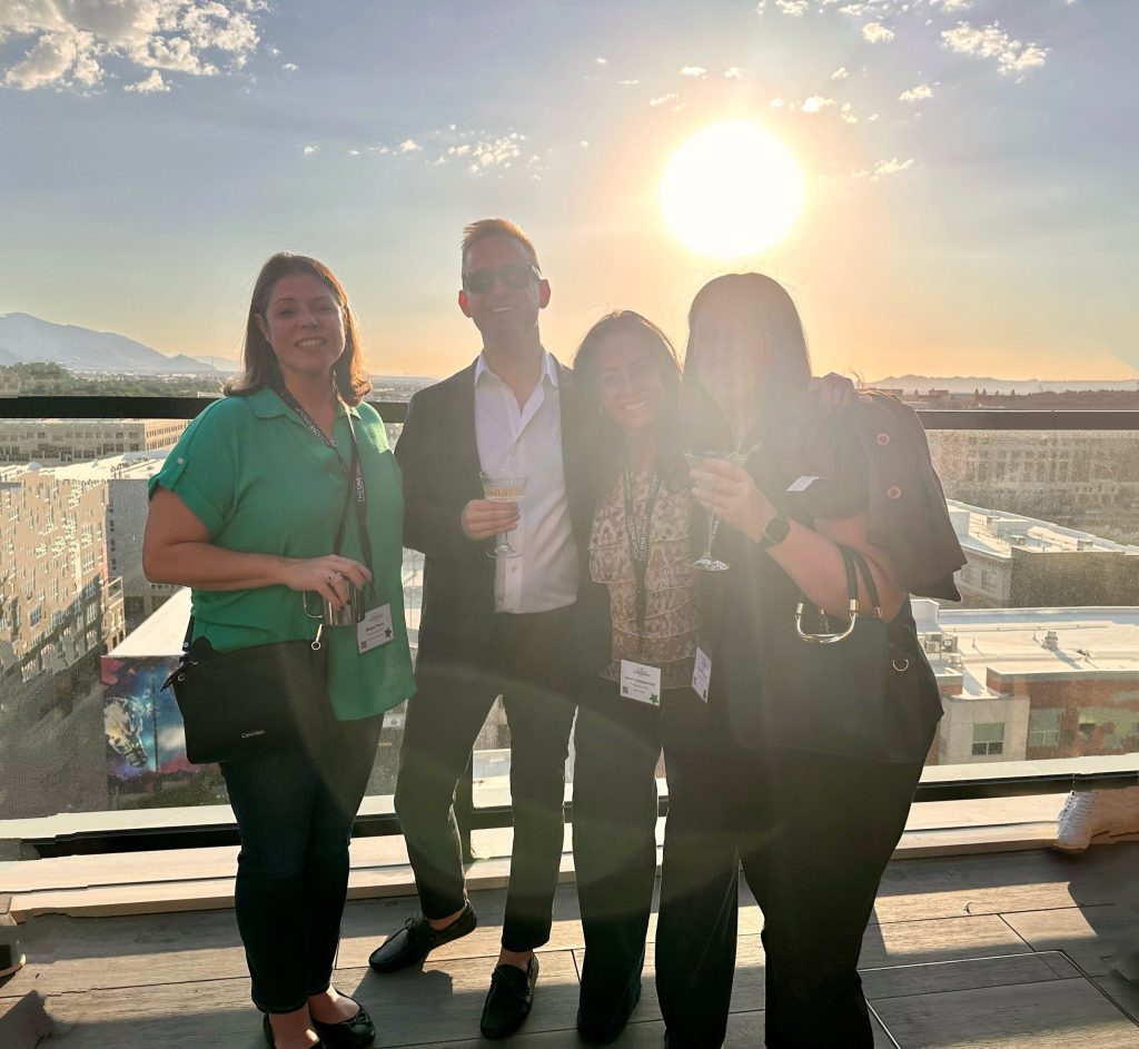 Four people smiling and holding drinks on a rooftop at sunset, with a cityscape and mountains in the background.