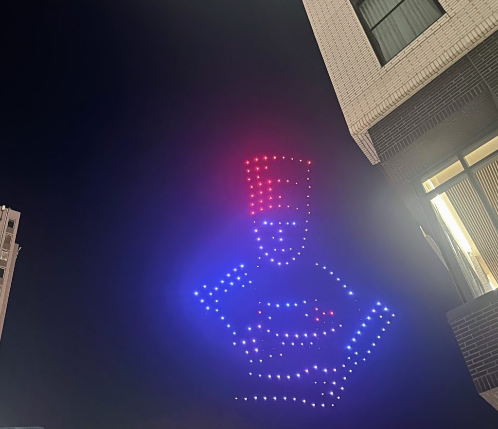 A night sky shows a formation of drones with lights creating the shape of a person in a hat. Part of a building is visible on the right.