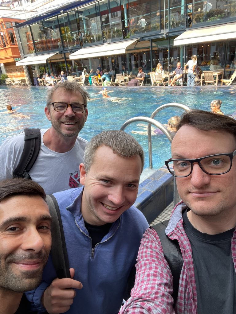 Four men smile near a swimming pool with people and seating areas in the background.